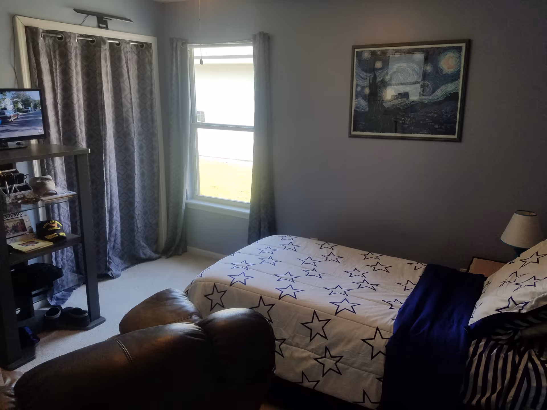 A furnished bedroom with a star-patterned bedspread, window, drapes, and a leather armchair.