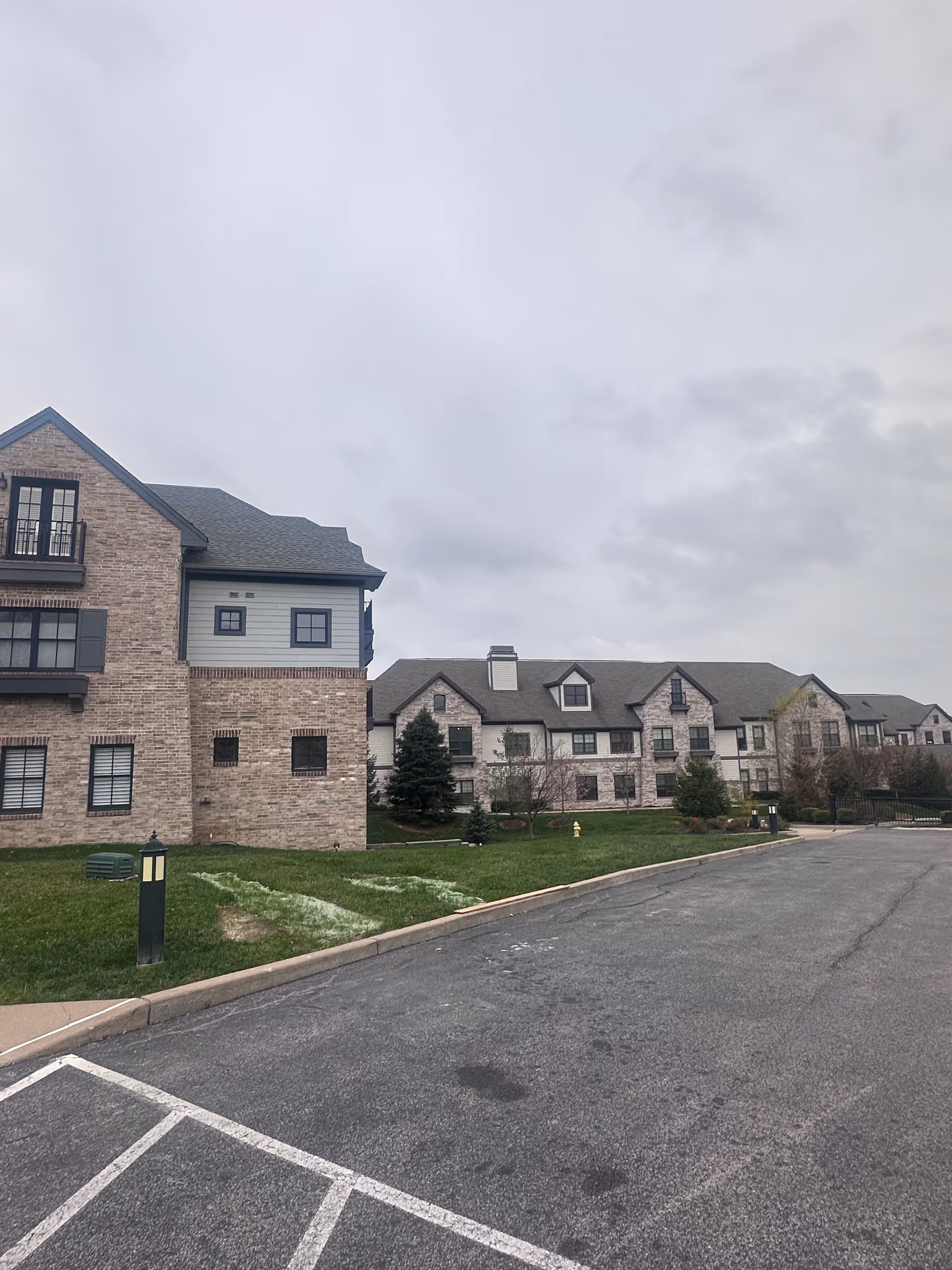 Exterior view of a multi-story brick and siding senior living building with a driveway and lawn under a cloudy sky.
