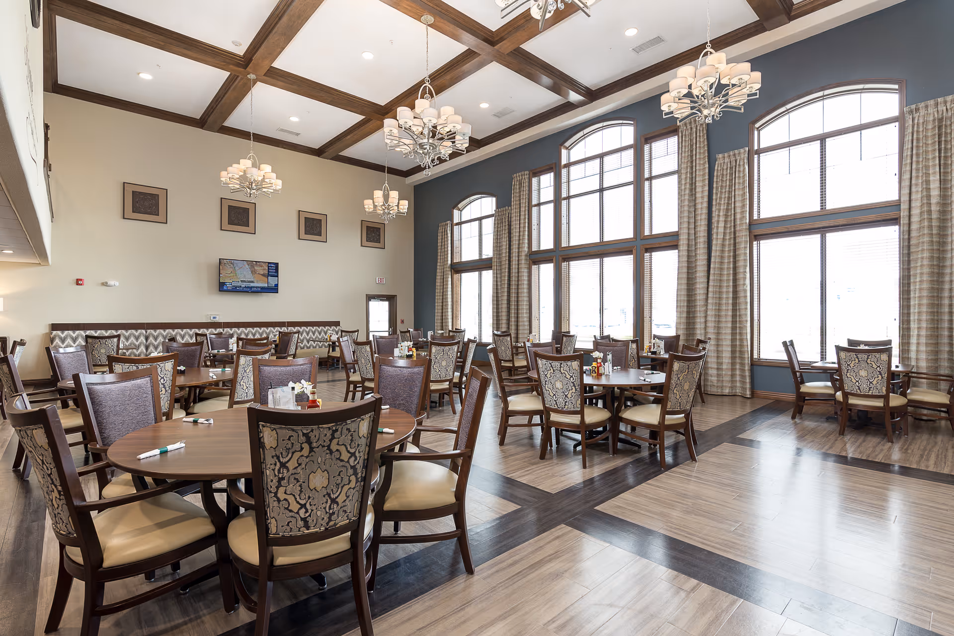 Spacious dining room with round tables, upholstered chairs, chandeliers, and large arched windows.