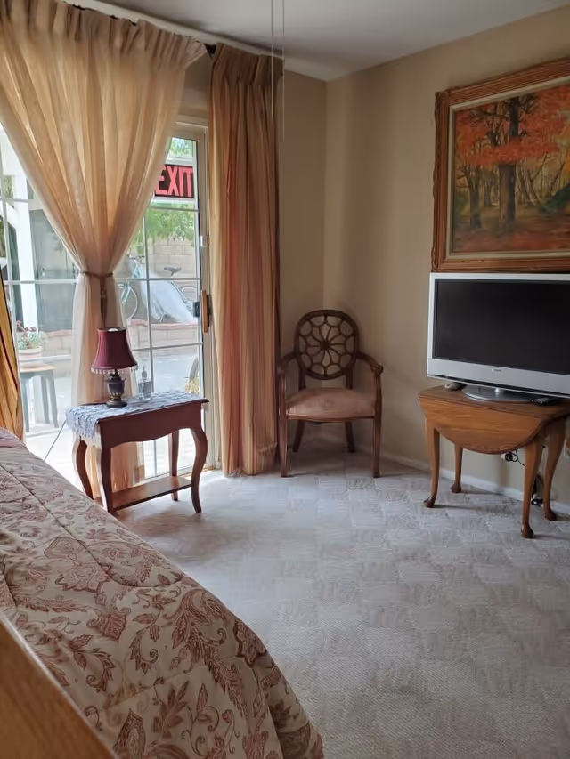 A cozy room with beige walls and carpet, featuring a bed with a patterned bedspread, a small wooden side table with a red lamp, a wooden chair with a decorative backrest, a flat-screen TV on a wooden table, and a large framed painting of autumn trees on the wall. Sheer beige curtains cover a sliding glass door with an EXIT sign visible outside.
