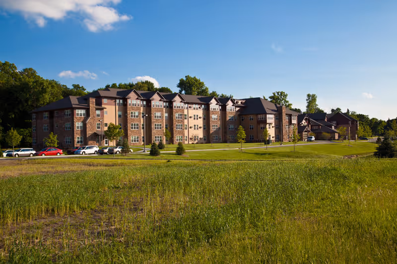 Multi-story brick senior living building with a green lawn, parked cars, and a blue sky.