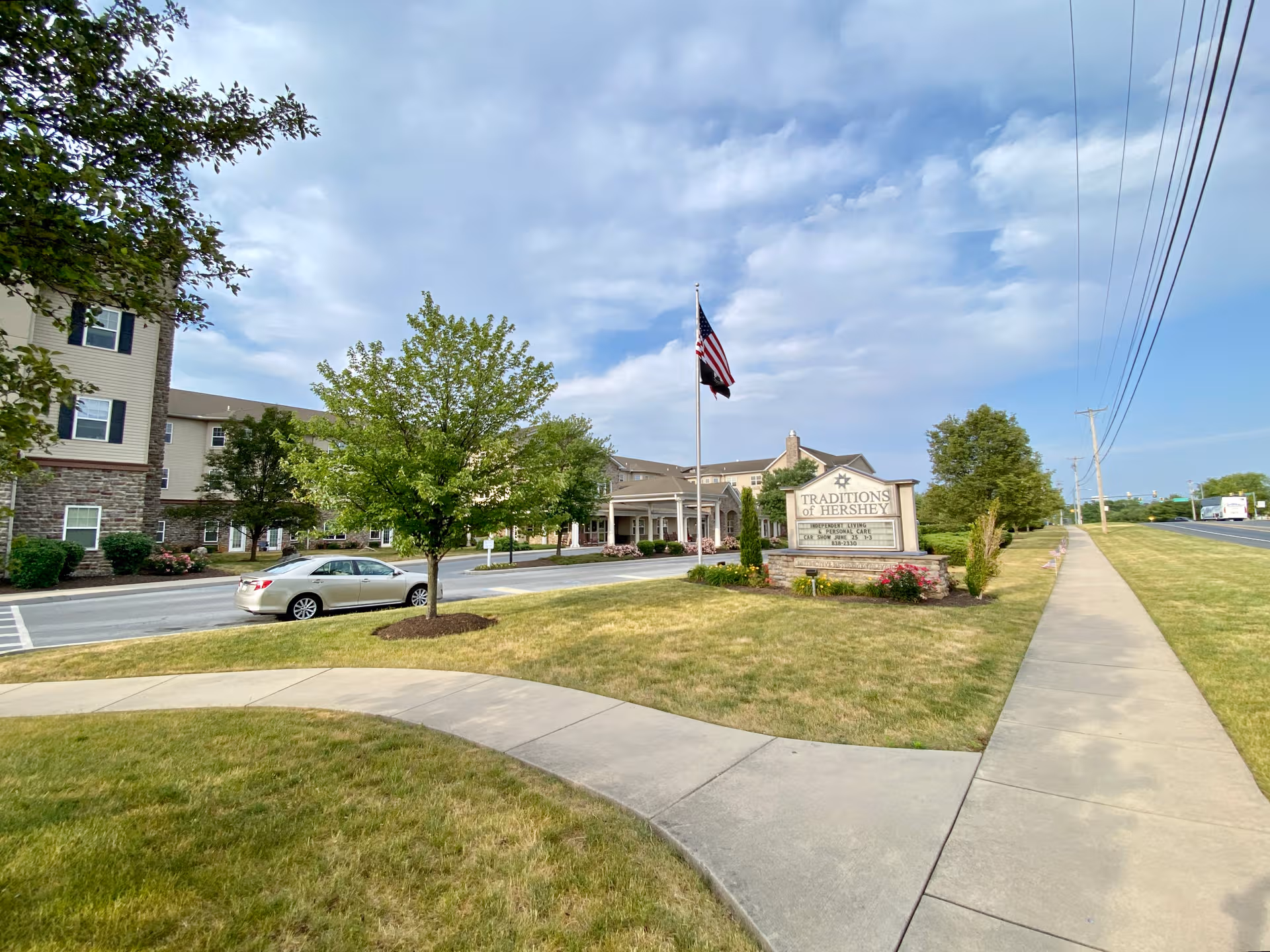 Exterior view of Traditions of Hershey senior living facility with entrance sign, flag, trees, and sidewalk.