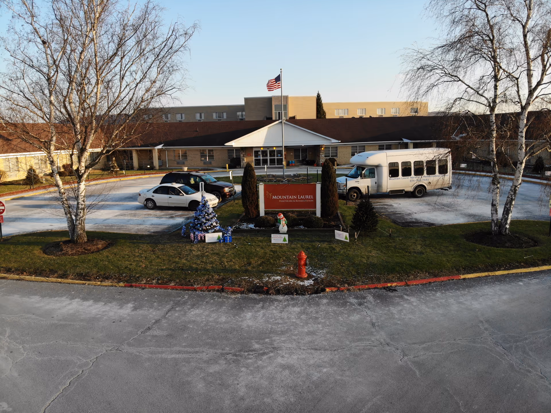Front exterior view of Mountain Laurel Healthcare and Rehabilitation Center with a circular driveway, parked vehicles including a white shuttle bus, an American flag on a flagpole, two leafless trees, a decorated Christmas tree with presents, and a red fire hydrant in the foreground.