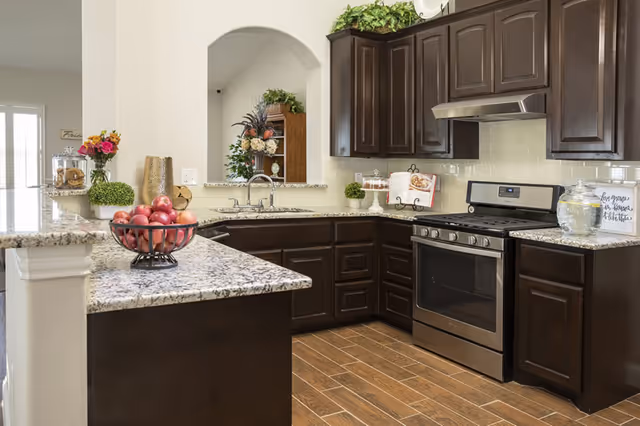 Modern kitchen with dark wooden cabinets, granite countertops, a stainless steel gas stove, a sink under an arched opening, and decorative items including a bowl of red apples, flowers, and glass jars.