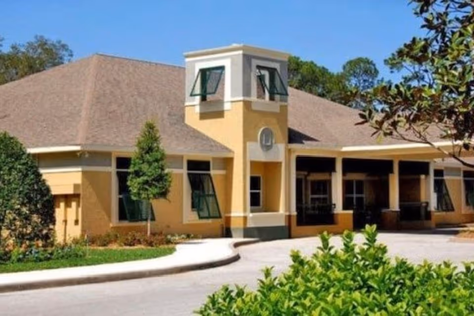 Exterior view of a single-story building with a beige and yellow facade, multiple windows with green awnings, a small tower-like structure in the center, surrounded by greenery and trees under a clear blue sky.