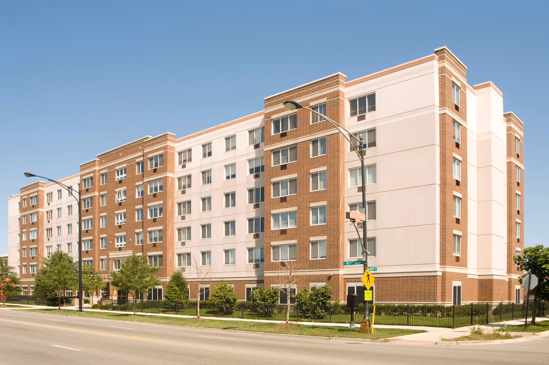 Exterior view of a multi-story senior living facility building with brick and light-colored paneling, surrounded by small trees and a sidewalk under a clear blue sky.