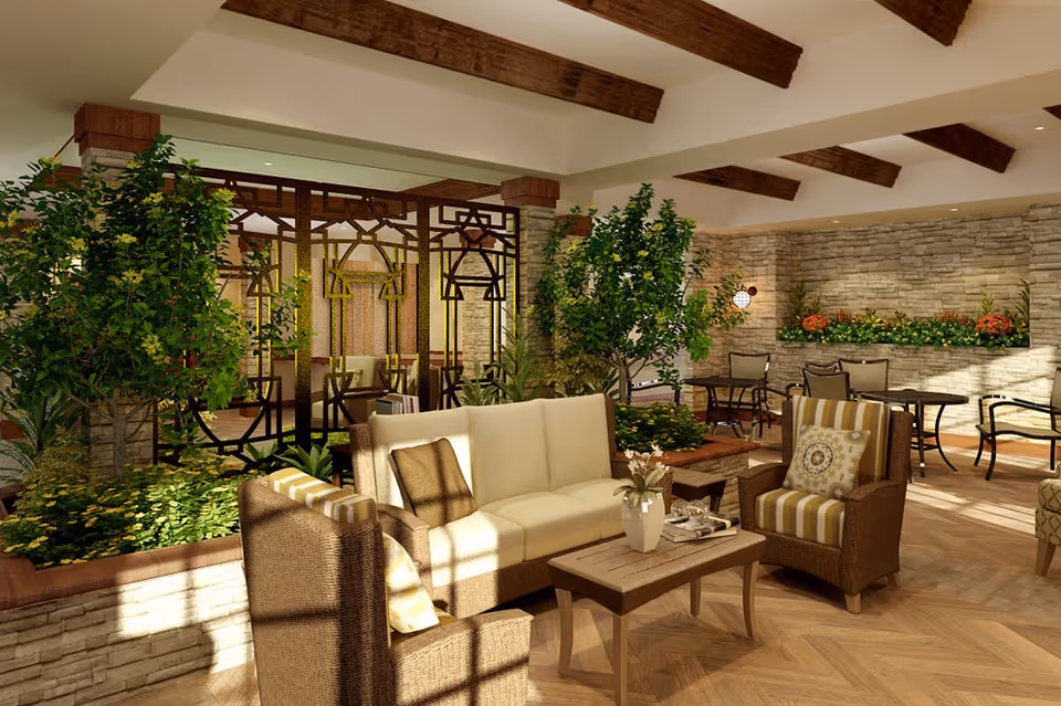 A bright and spacious indoor lounge area with comfortable wicker furniture including a cream-colored sofa and striped armchairs around a wooden coffee table. The room features wooden ceiling beams, stone walls, lush green plants, and decorative metal partitions, creating a warm and inviting atmosphere.