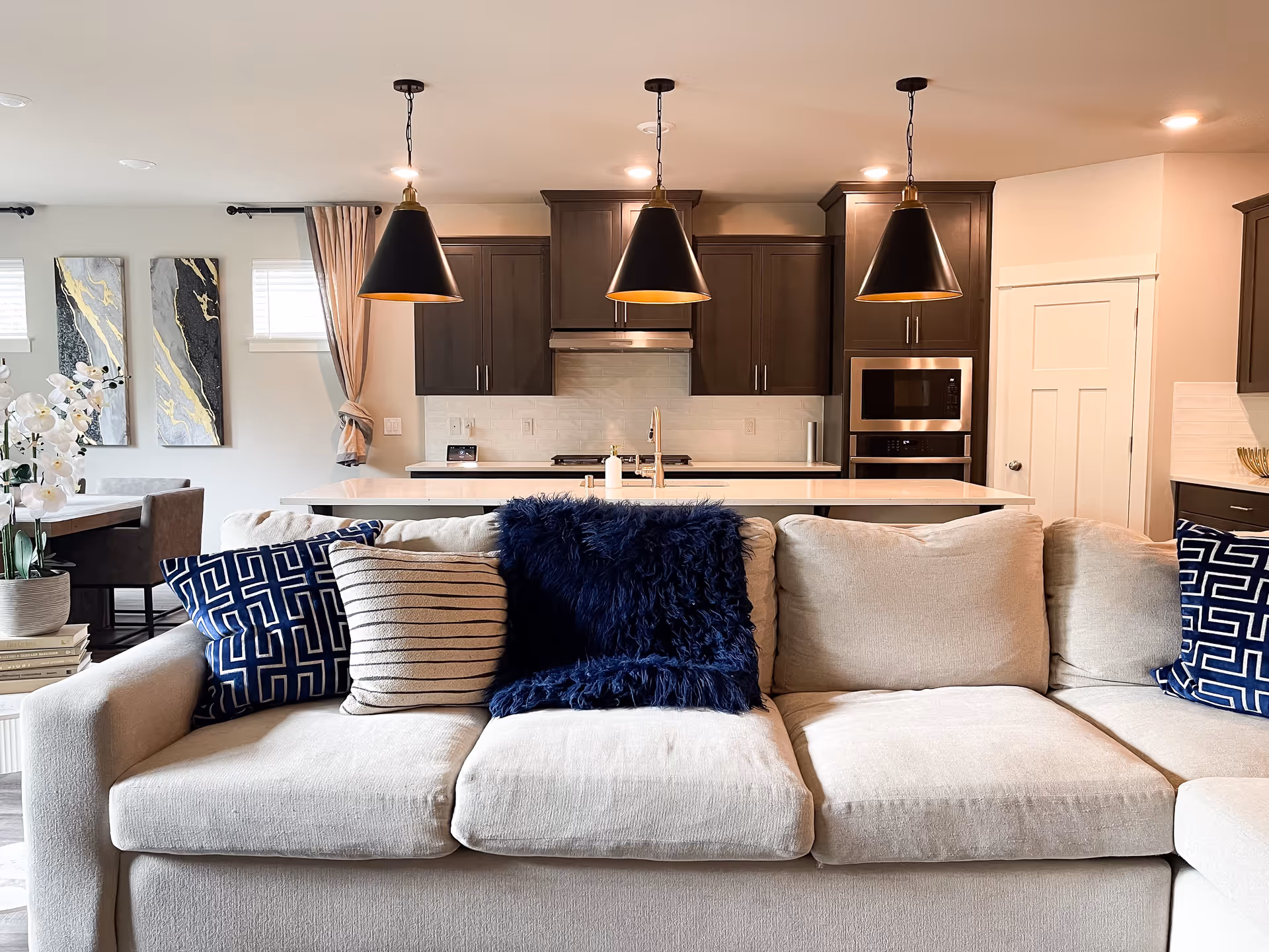 Open-plan living area with a beige sofa topped with blue and striped pillows in front of a modern kitchen island with three pendant lights.