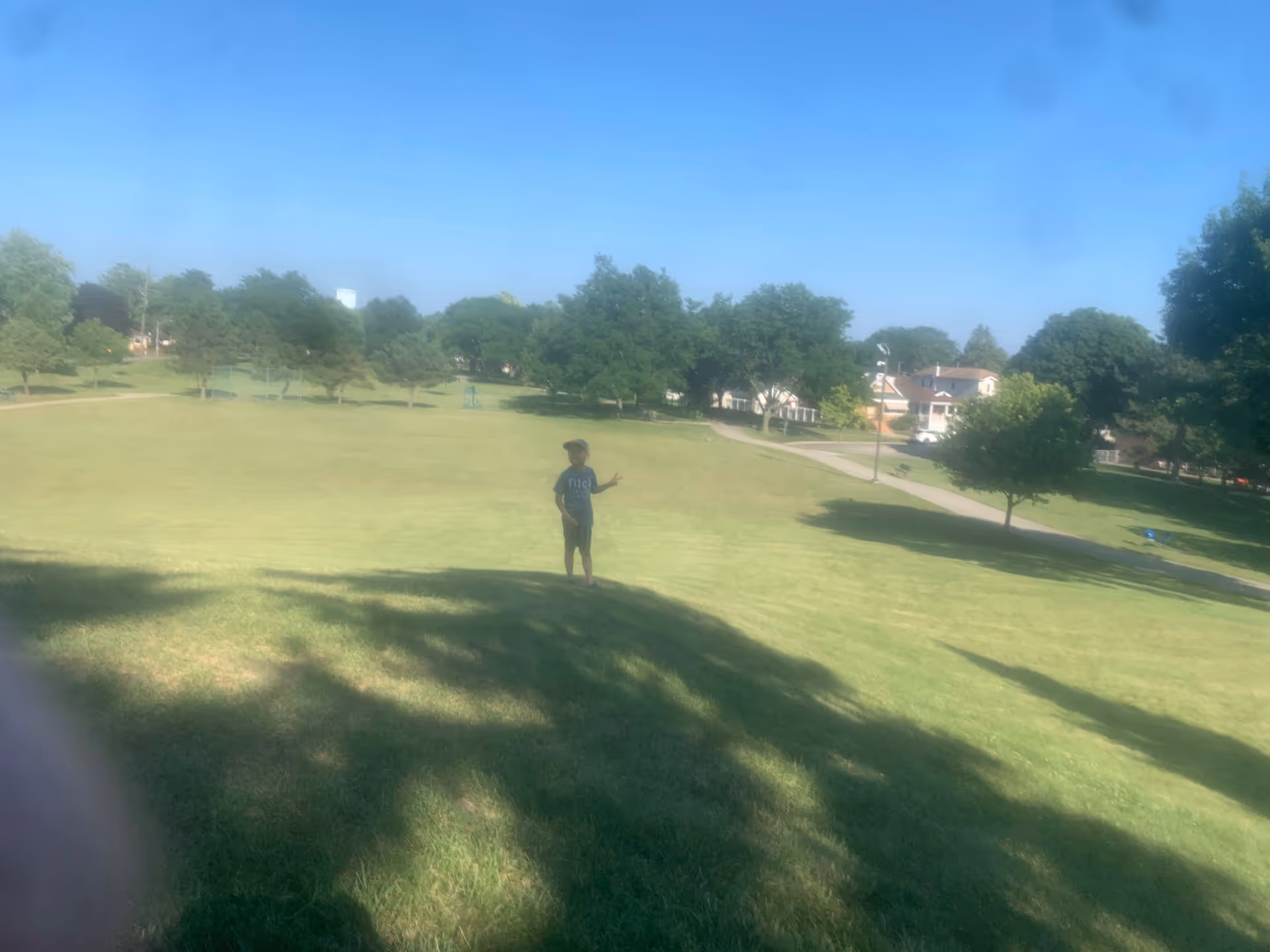 A person stands on a grassy park area with trees and houses in the background.