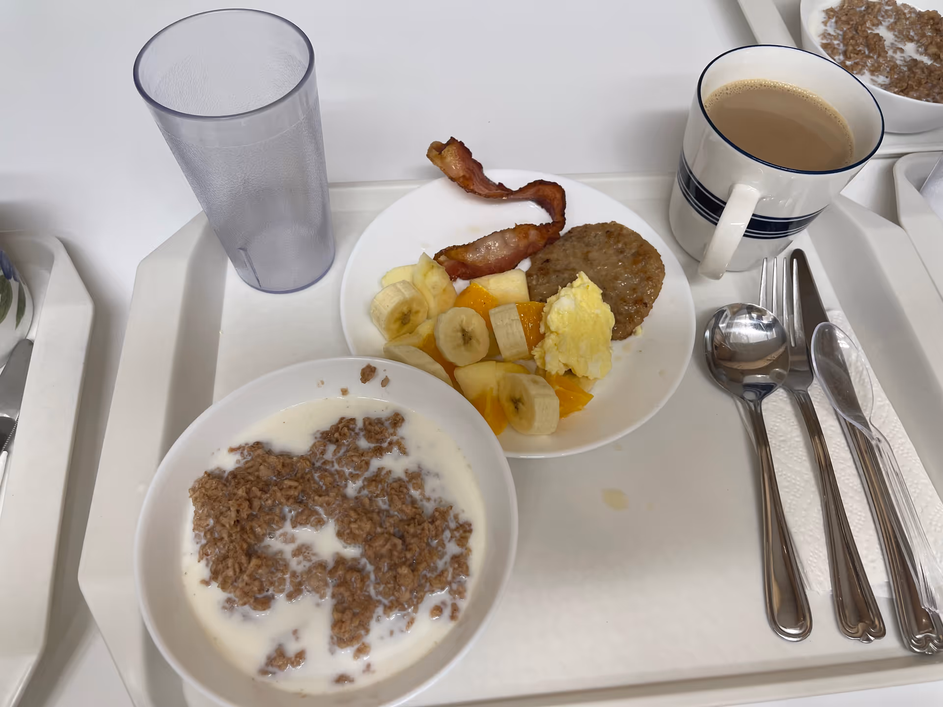 Breakfast tray with sausage, eggs, fruit, cereal, and coffee on a white dining table.