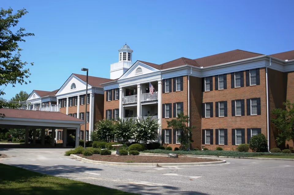 Front view of a three-story brick senior living building with a covered entrance, balconies, an American flag, and landscaped driveway under a clear blue sky.