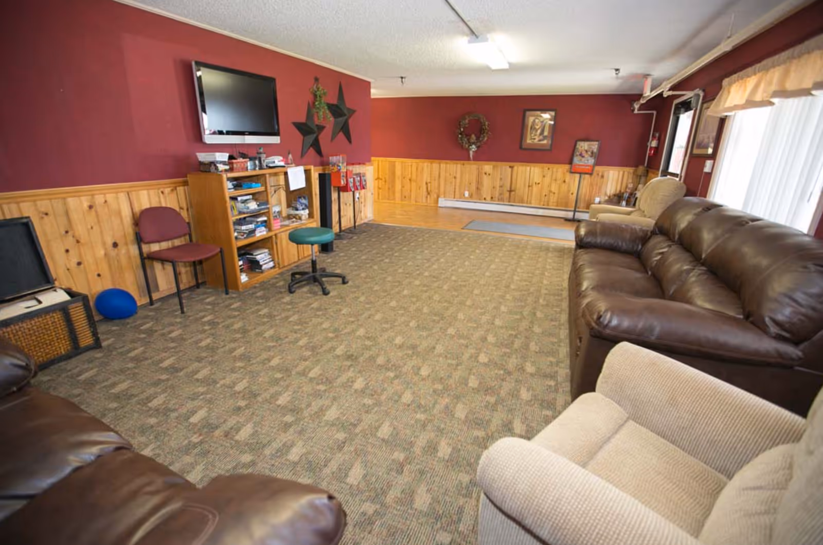 A spacious living room with maroon walls and wood paneling. The room features a large brown leather sofa, a beige armchair, a maroon chair, a green swivel stool, and a wooden bookshelf filled with books and games. A flat-screen TV is mounted on the wall above the bookshelf. The room has carpeted flooring and large windows with sheer curtains allowing natural light to enter.