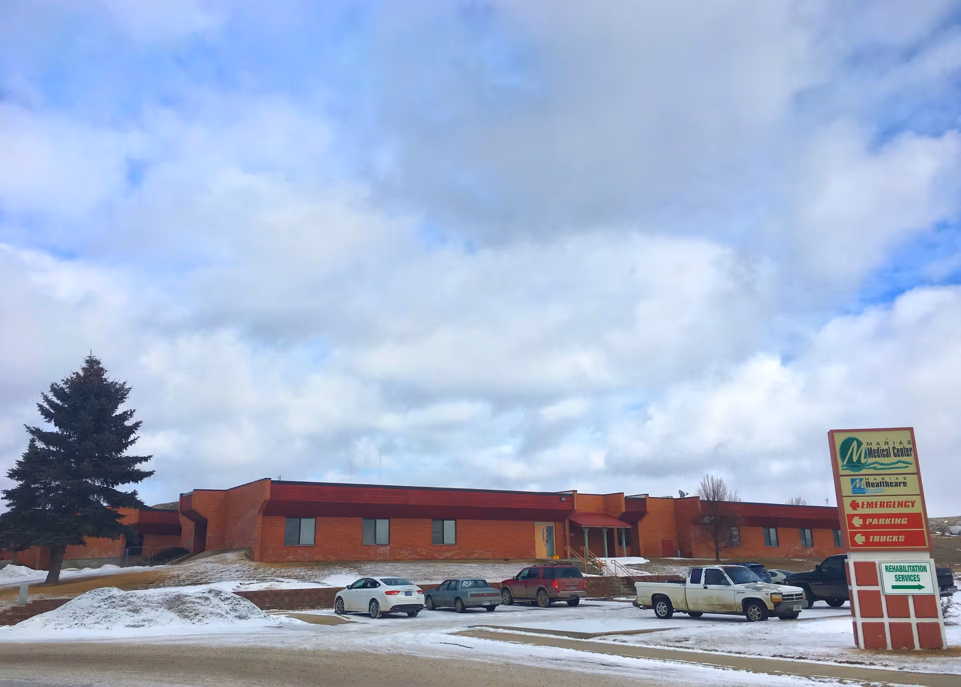 Exterior view of a red brick senior care facility with parking and a cloudy sky.