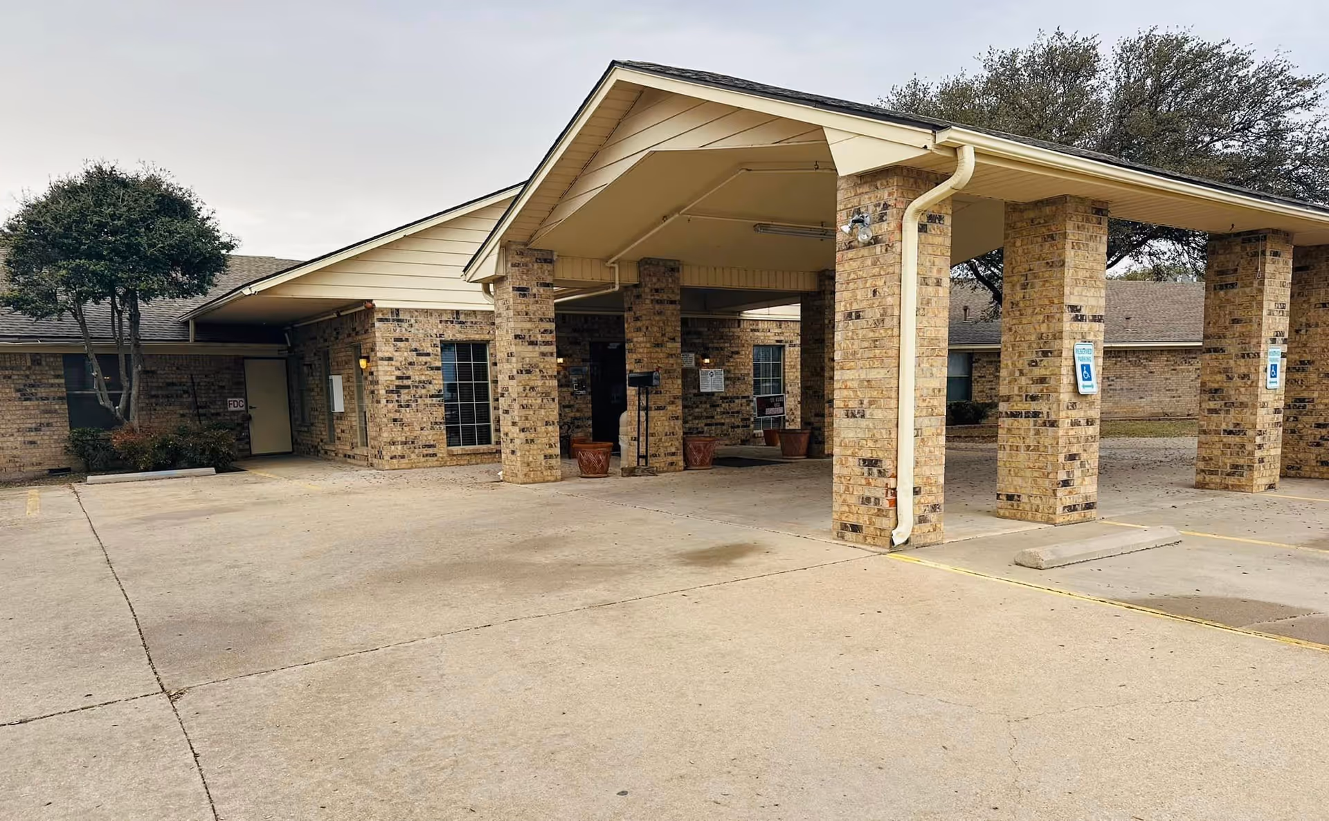 Exterior view of Ballinger Healthcare & Rehab Center showing a covered entrance with brick pillars and a concrete driveway. There are handicap parking signs on the pillars and some trees and shrubs around the building.