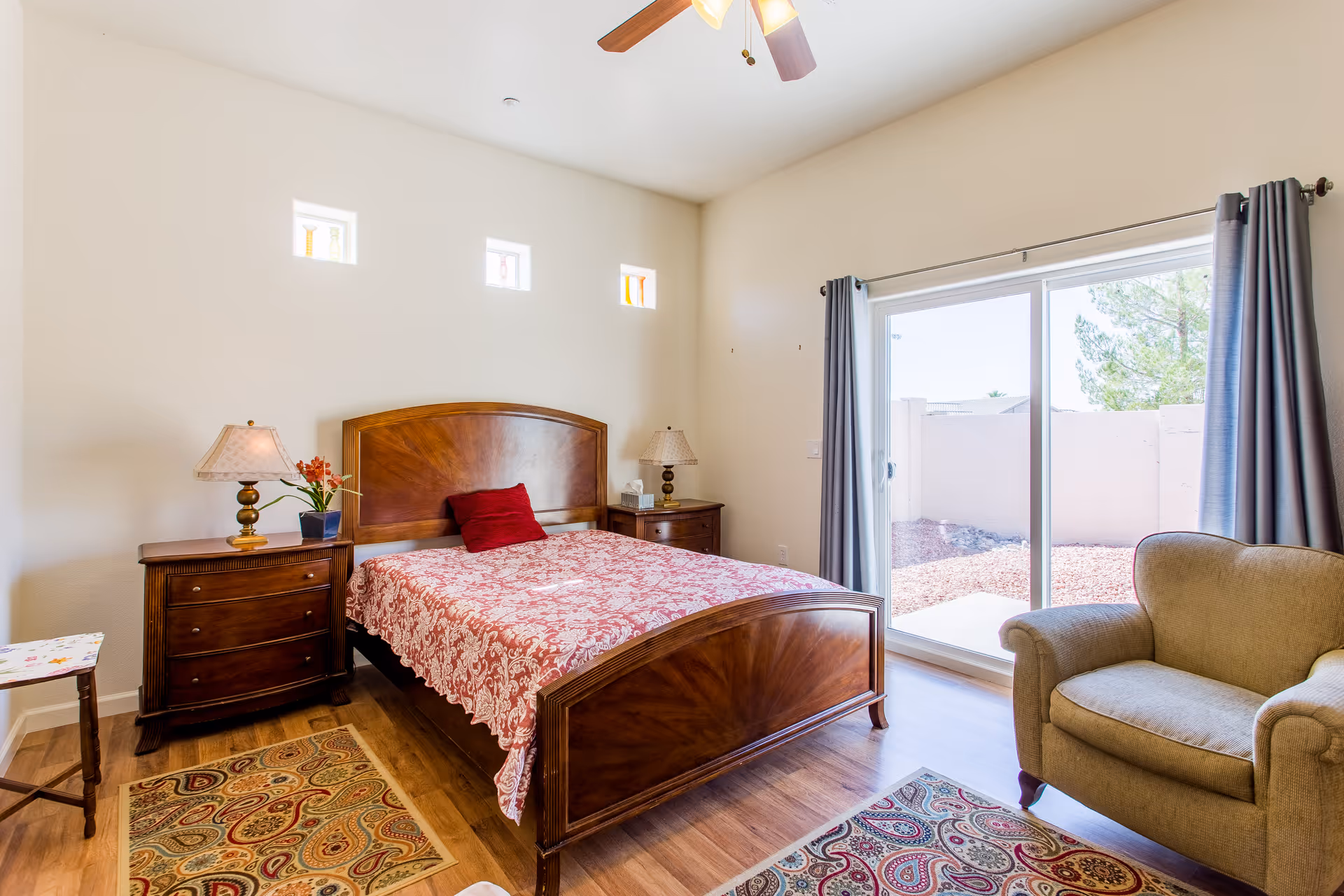 A bright bedroom with a wooden bed featuring a red patterned bedspread and a red pillow. There are two wooden nightstands with lamps and a small plant on one side. The room has three small square windows near the ceiling and a large sliding glass door with gray curtains leading to an outdoor area. A beige armchair and two patterned rugs are also visible on the wooden floor.