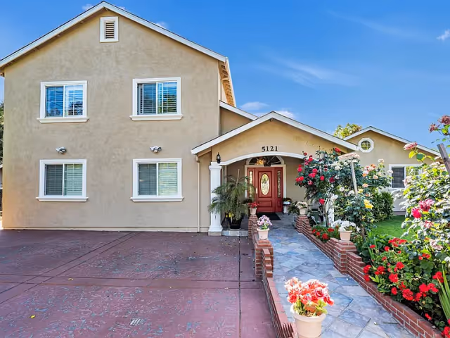 Two-story beige building with address 5121, a red front door, driveway and a flower-lined tiled walkway.