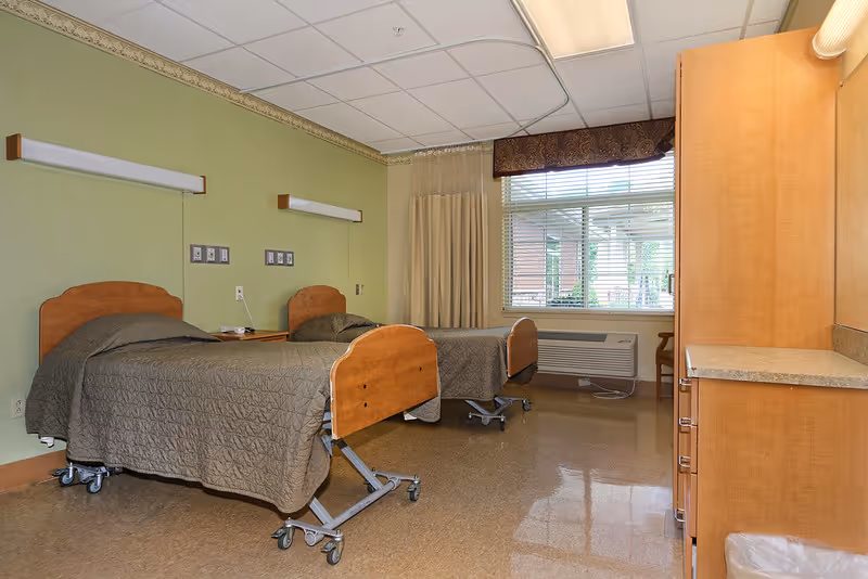 Two hospital-style beds in a green-walled senior living bedroom with curtains.