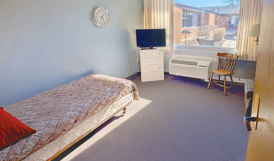 A simple furnished bedroom with a single bed, dresser, TV, clock, and window view.