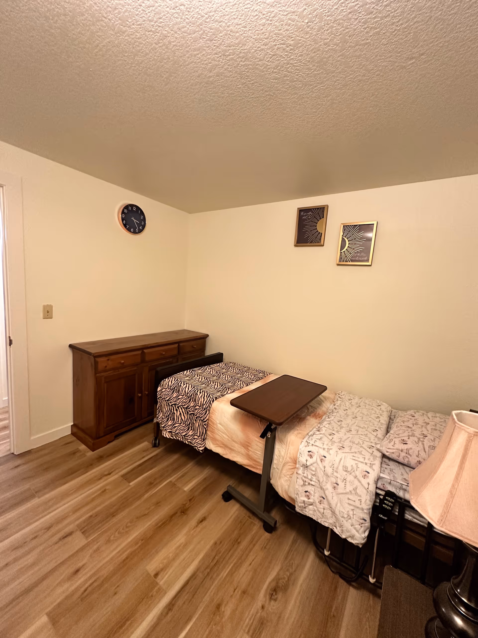 Two single beds with an overbed table, dresser, clock, and wall art in a small bedroom.