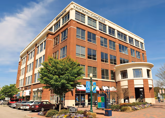 Brick multi-story senior living facility exterior with storefronts, signage, and parked cars along street