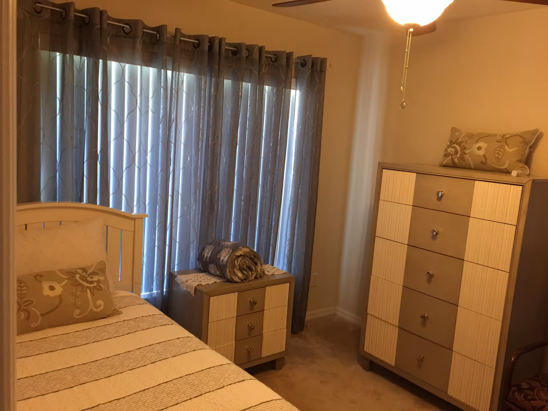 A cozy bedroom with a single bed featuring a striped bedspread and a floral pillow. Next to the bed is a small nightstand with a rolled blanket on top. A tall dresser with multiple drawers and a decorative pillow on top is positioned against the wall. The room has beige walls, carpeted floor, and a window covered with patterned blue curtains.