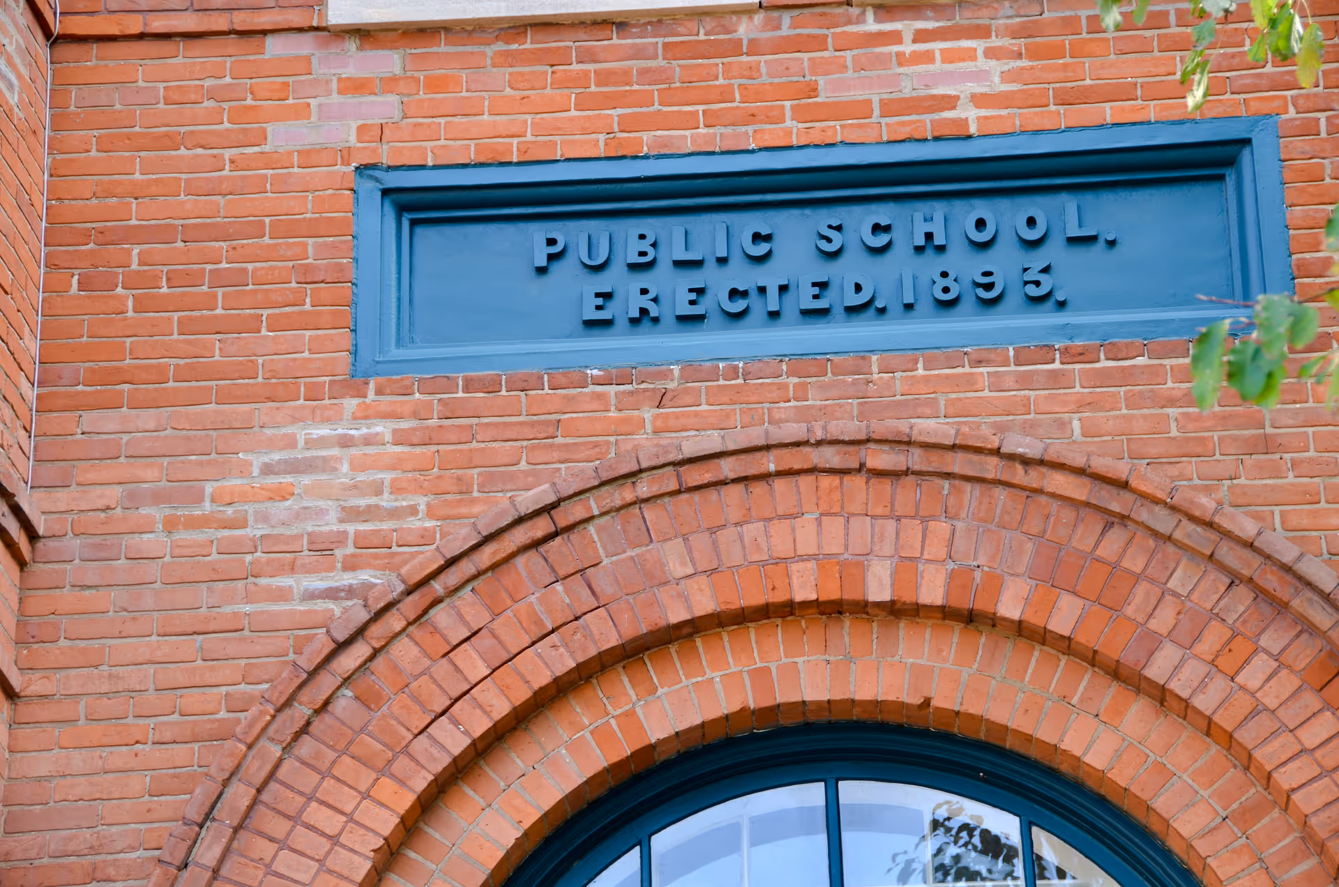 Close-up view of a red brick building facade with an arched window below a blue plaque that reads 'PUBLIC SCHOOL, ERECTED, 1893.'