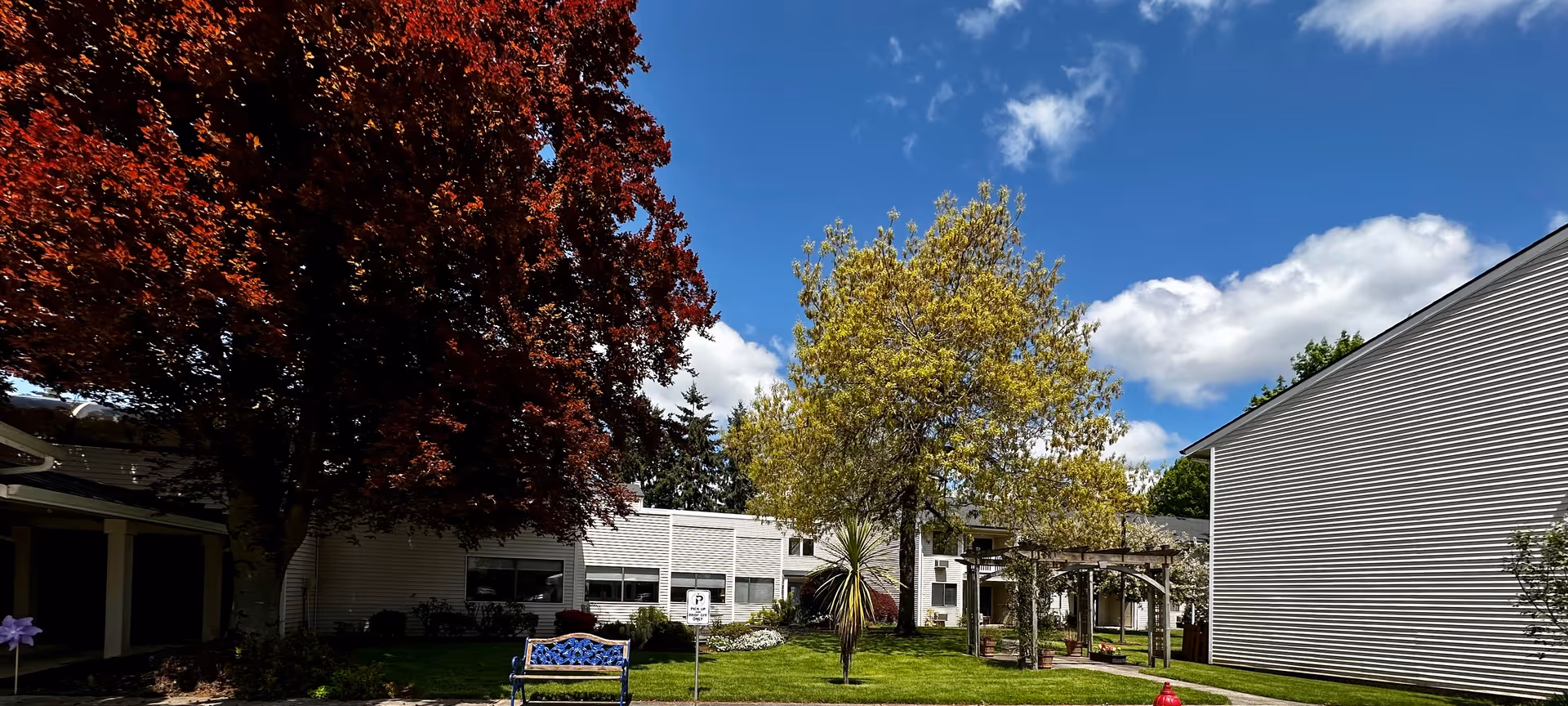 A sunny courtyard with trees, a blue bench, grassy lawn, and surrounding retirement facility buildings under a blue sky.