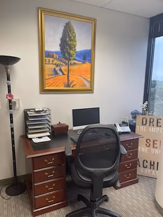 Office desk with computer, chair, lamp, and framed landscape painting on wall.