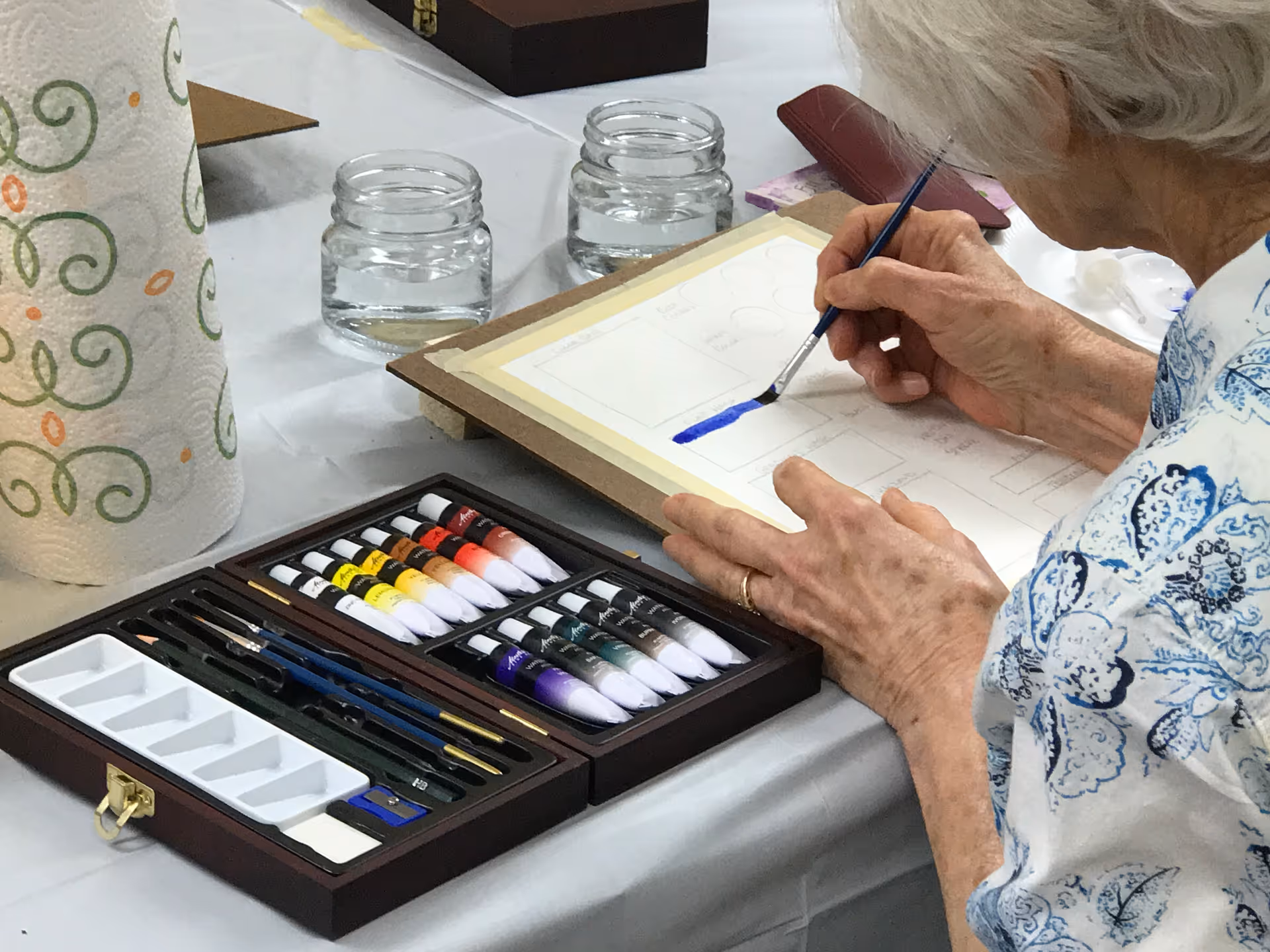 An elderly person with white hair is painting with a brush on a piece of paper taped to a board. There is a set of watercolor paints, brushes, and two glass jars of water on the table. A roll of paper towels is also visible on the left side of the table.