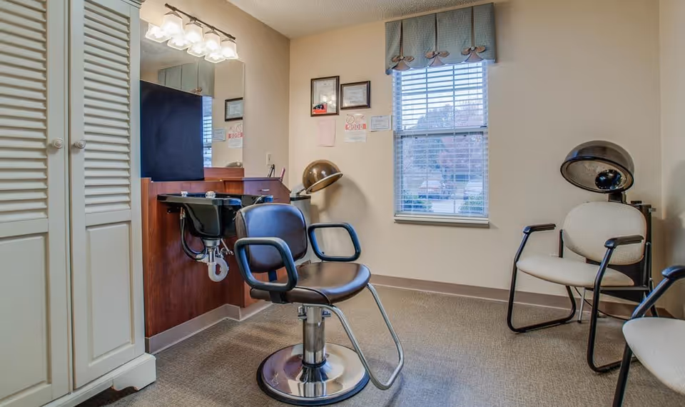 Small hair salon room with a styling chair, shampoo sink, hooded hair dryer and waiting chairs by a window.