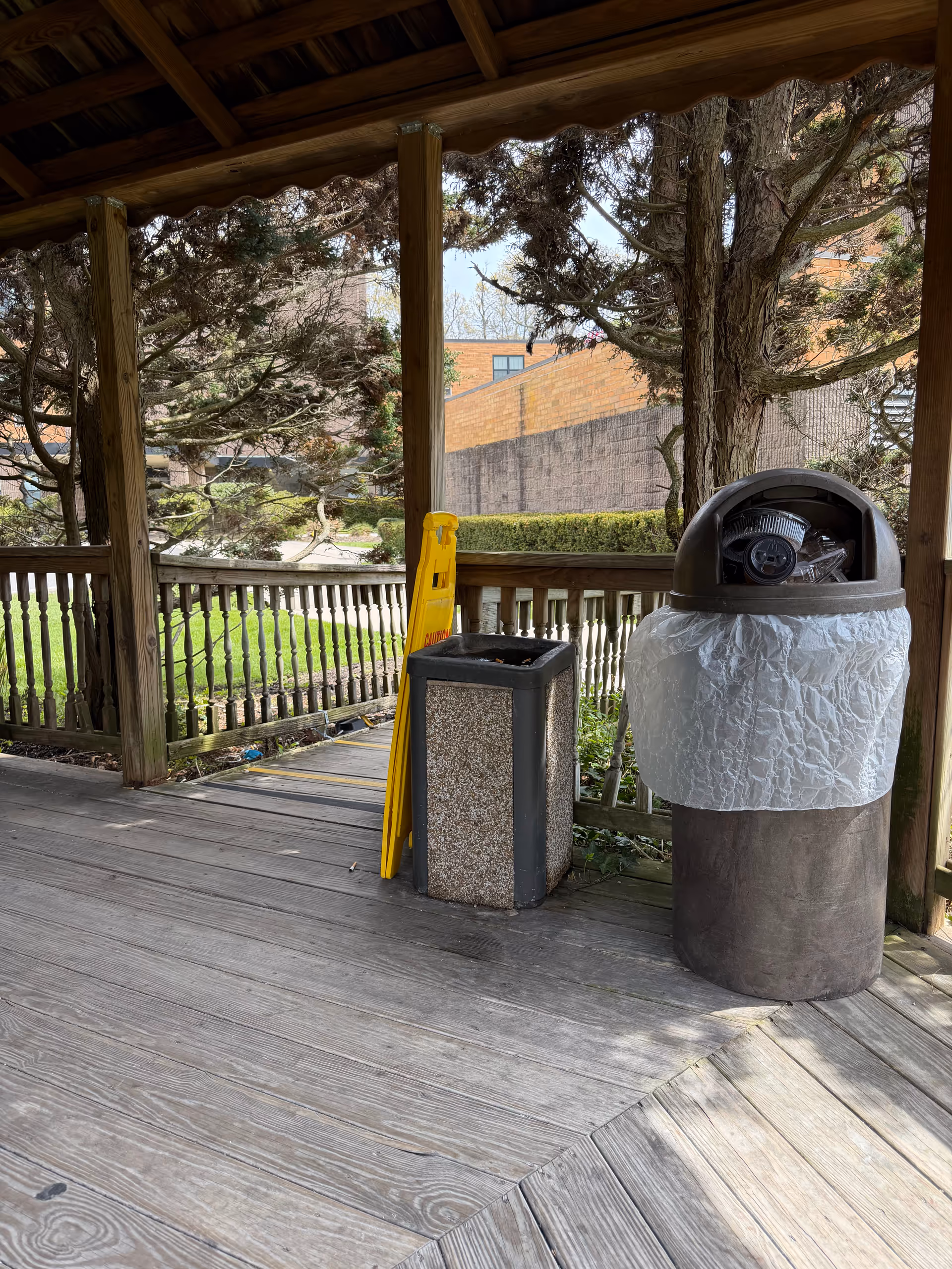 Covered wooden porch at Surge Rehabilitation and Nursing with railing, trees, caution sign, and trash bin.
