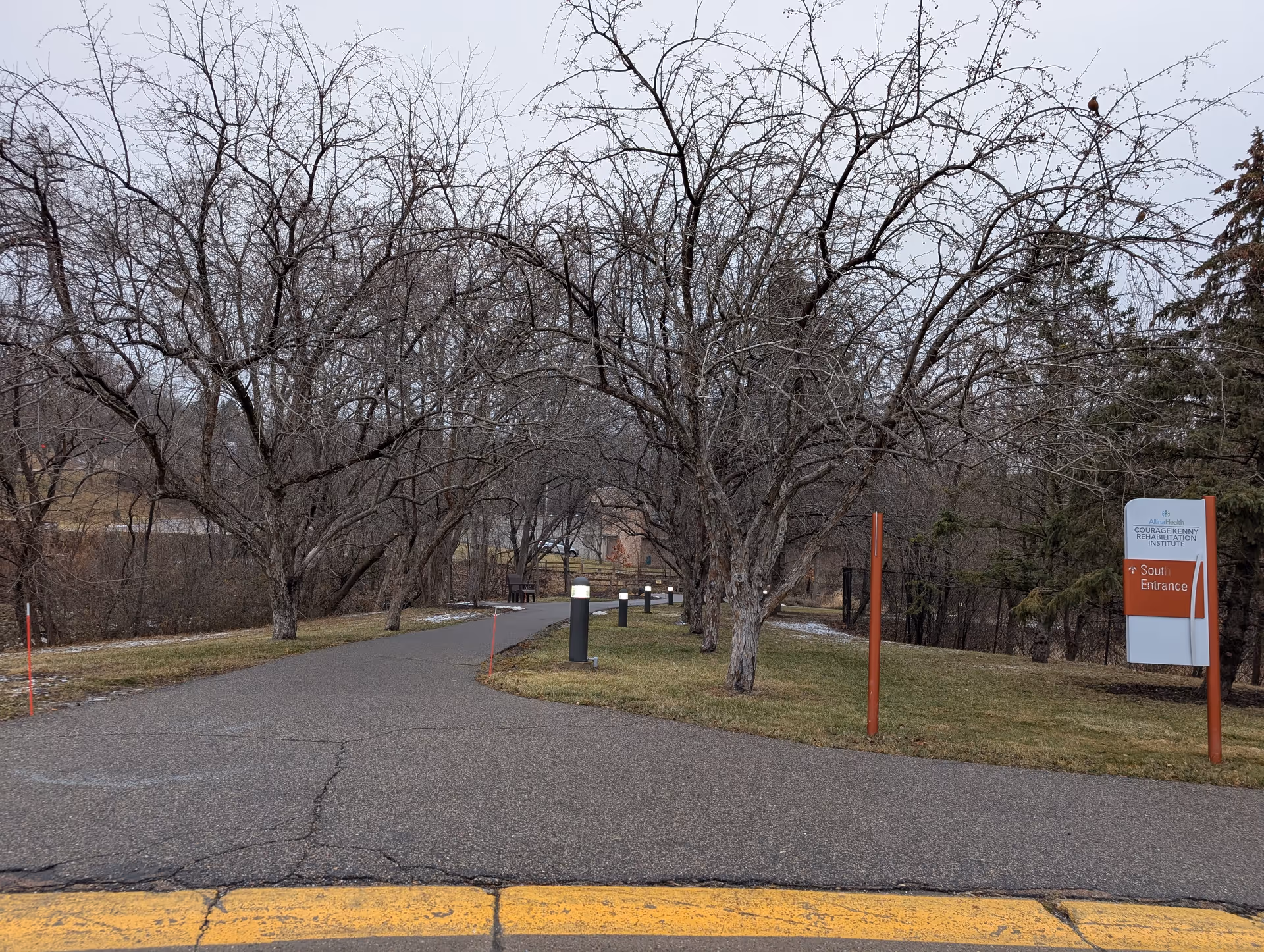 A paved pathway lined with leafless trees on both sides leading into a wooded area. There are small black lamp posts along the path and a sign on the right side that reads 'Allina Health Courage Kenny Rehabilitation Institute South Entrance'. The sky is overcast and there is some sparse snow on the grass.