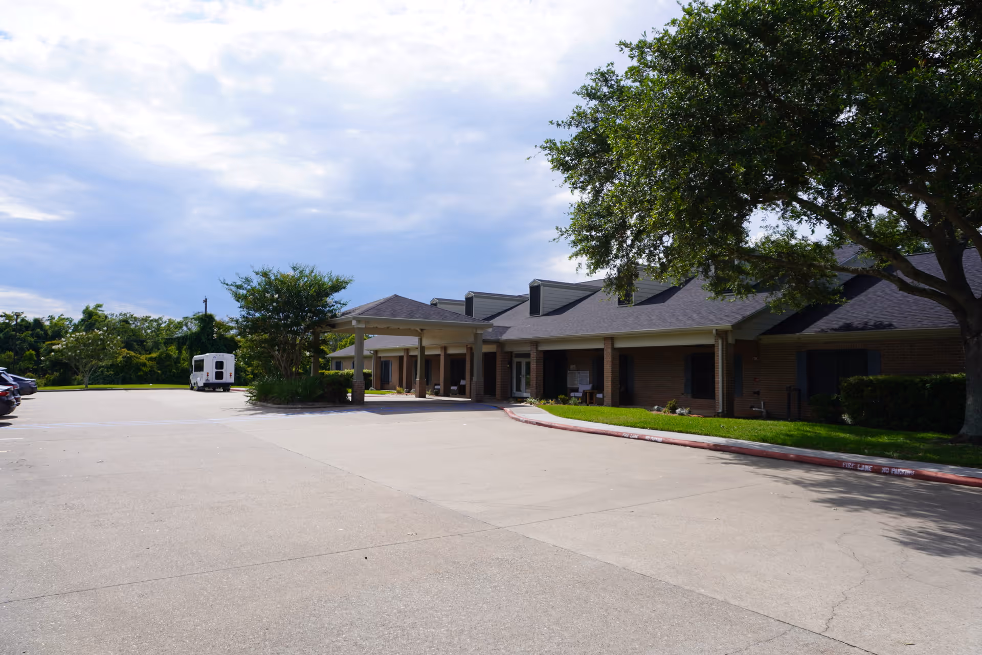 Exterior view of a single-story brick building with a covered entrance and a parking lot in front. There are trees and greenery around the building under a partly cloudy sky.