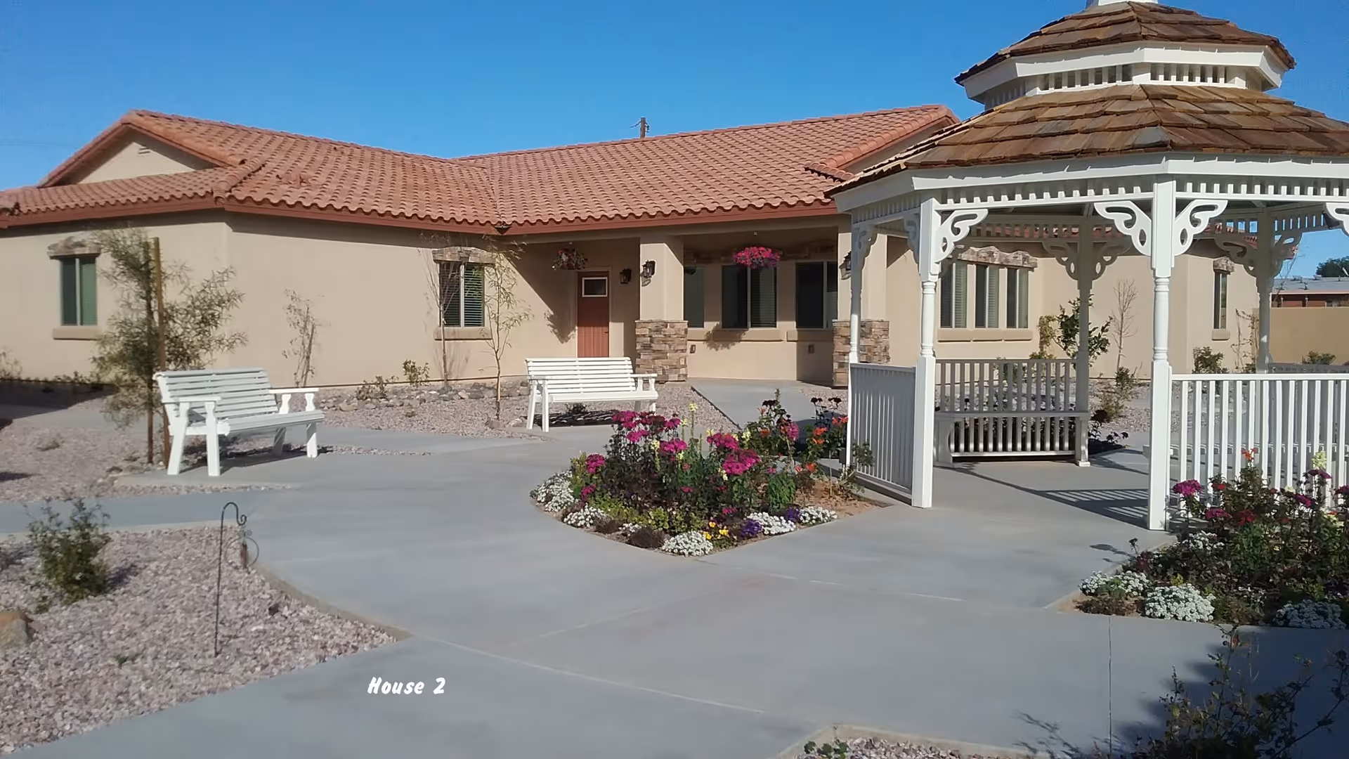 Courtyard with a white gazebo, flower beds and benches in front of a single-story stucco building with a tiled roof.