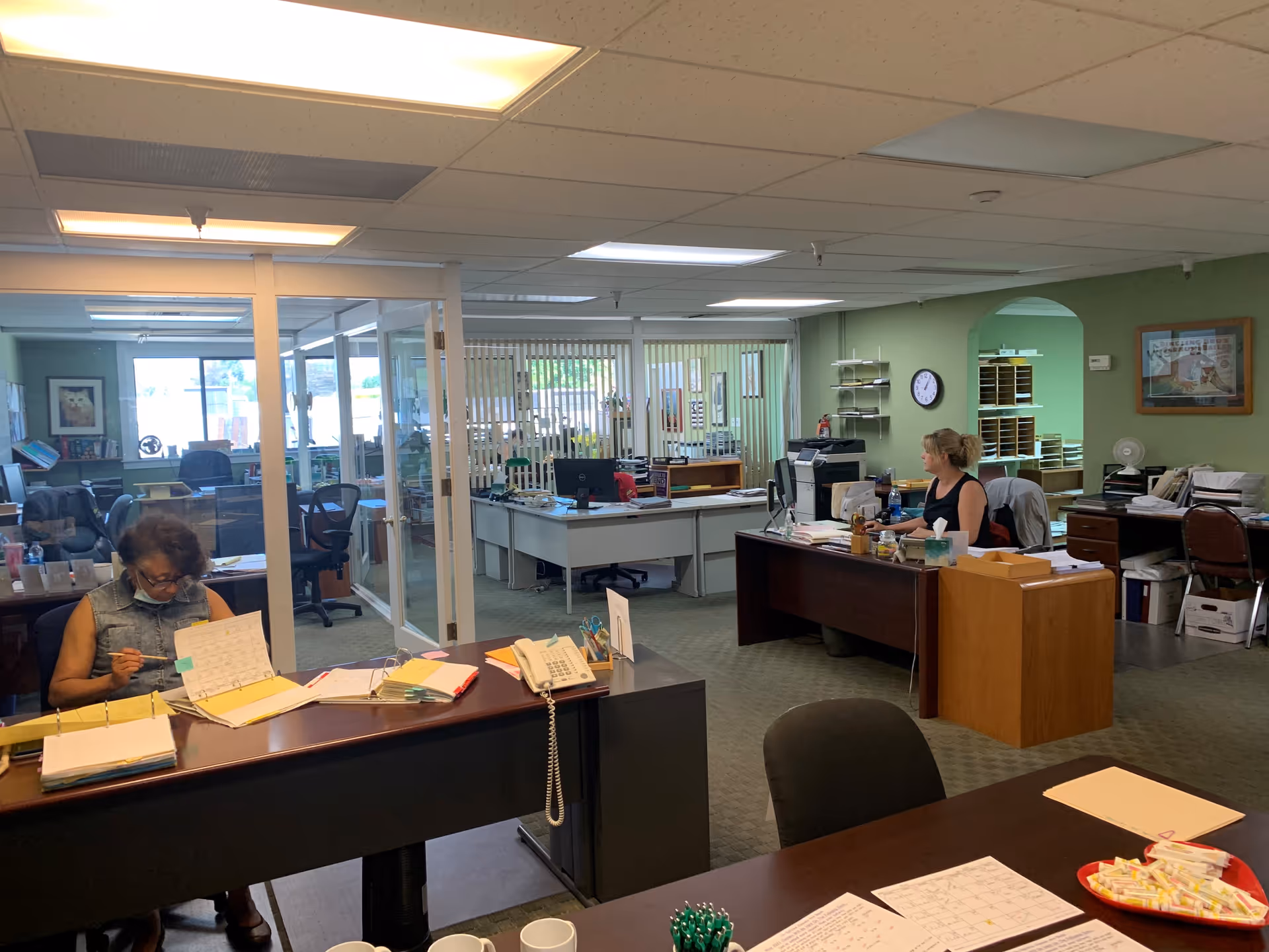 Two staff members work at desks in a busy office with glass partitions.