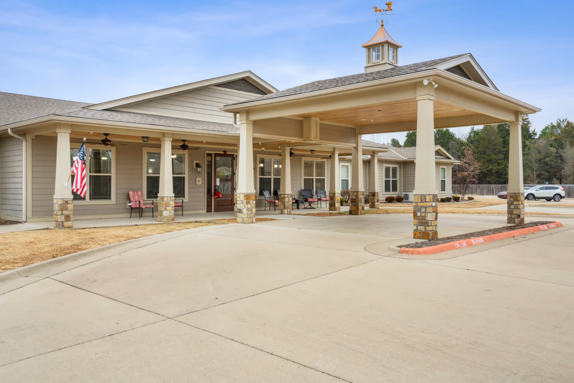 Exterior view of StoneBridge Senior Living - Russellville showing the entrance with a covered driveway supported by columns with stone bases. There are chairs on the porch, an American flag, and a weather vane on top of the roof. The surrounding area includes a parking lot and some trees in the background.