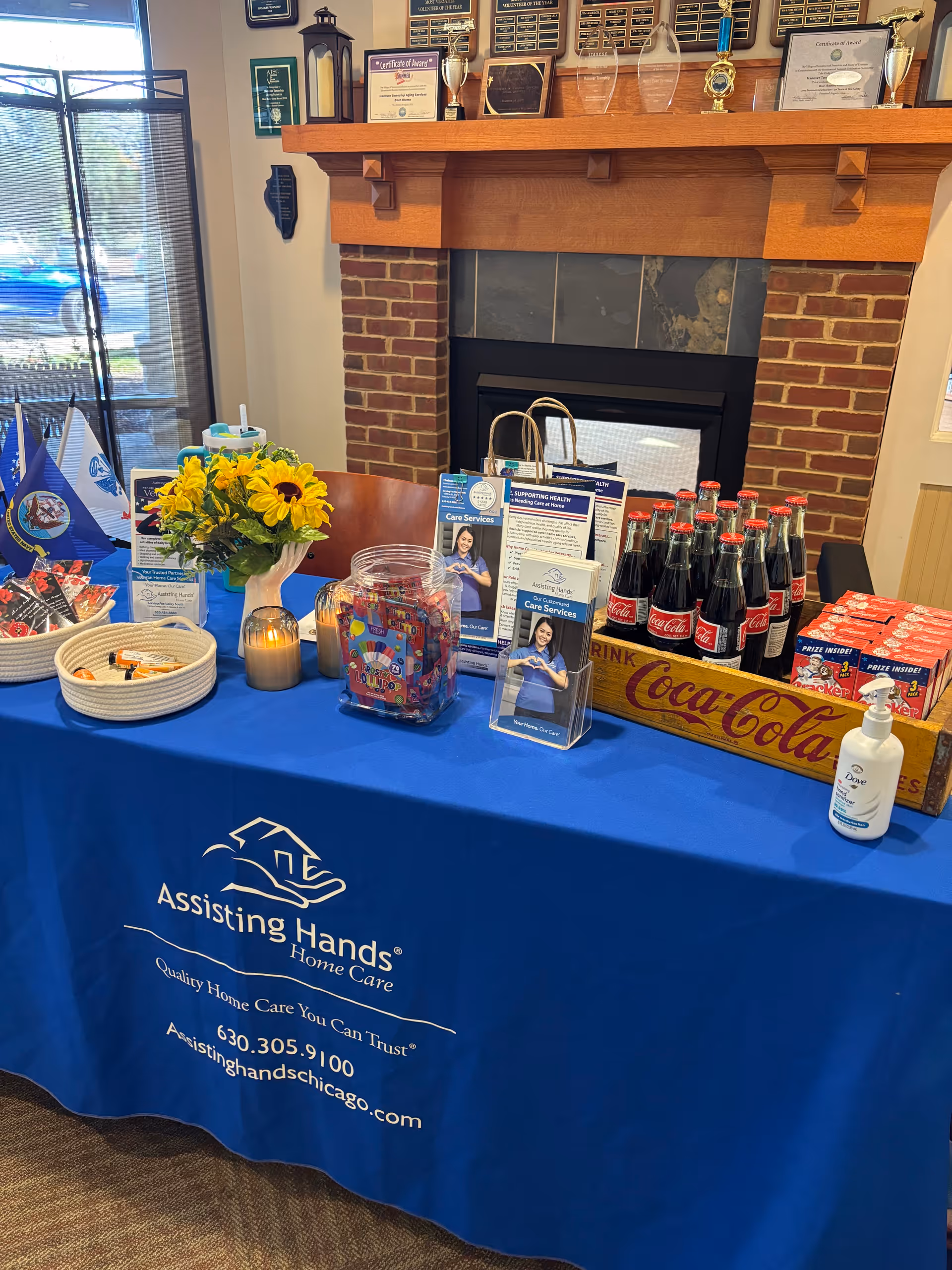 Blue promotional table at an interior reception area with brochures, flowers, snacks, and Coca-Cola bottles.