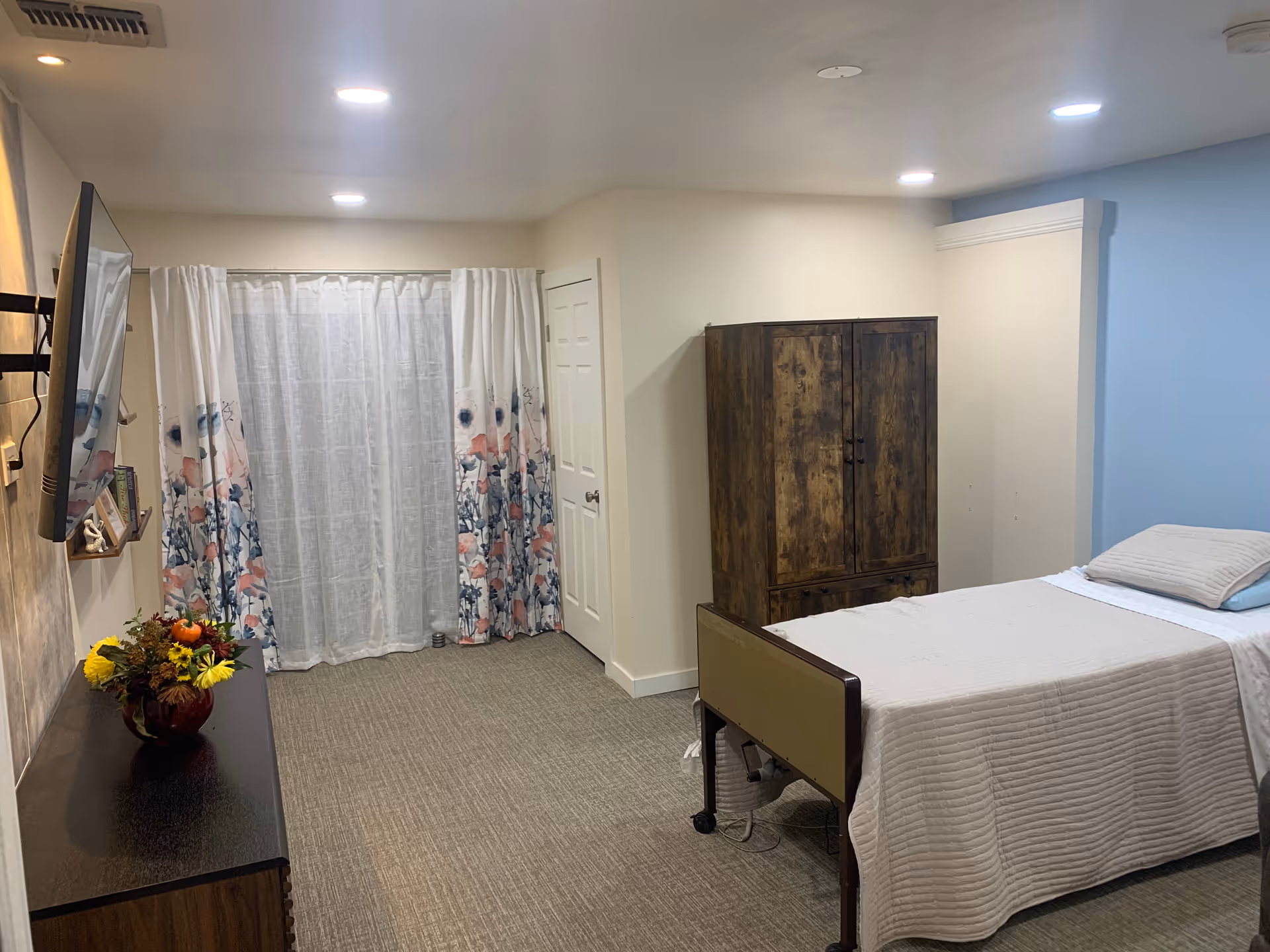 A simple, clean bedroom in a senior living facility with a single bed covered in a beige quilt, a wooden wardrobe, a dresser with a vase of flowers, a wall-mounted TV, and floral curtains covering a window or door.