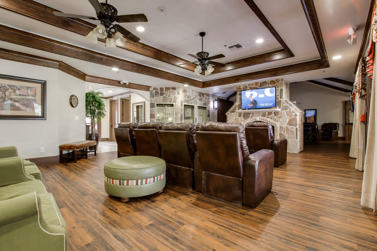 A spacious living room with wooden flooring and ceiling beams, featuring a row of brown leather recliners facing a stone fireplace with a mounted flat-screen TV. There are green upholstered chairs and a round green ottoman with a striped band in the foreground. The room is well-lit with recessed lighting and ceiling fans with lights. A framed picture and a potted plant are visible on the left wall.
