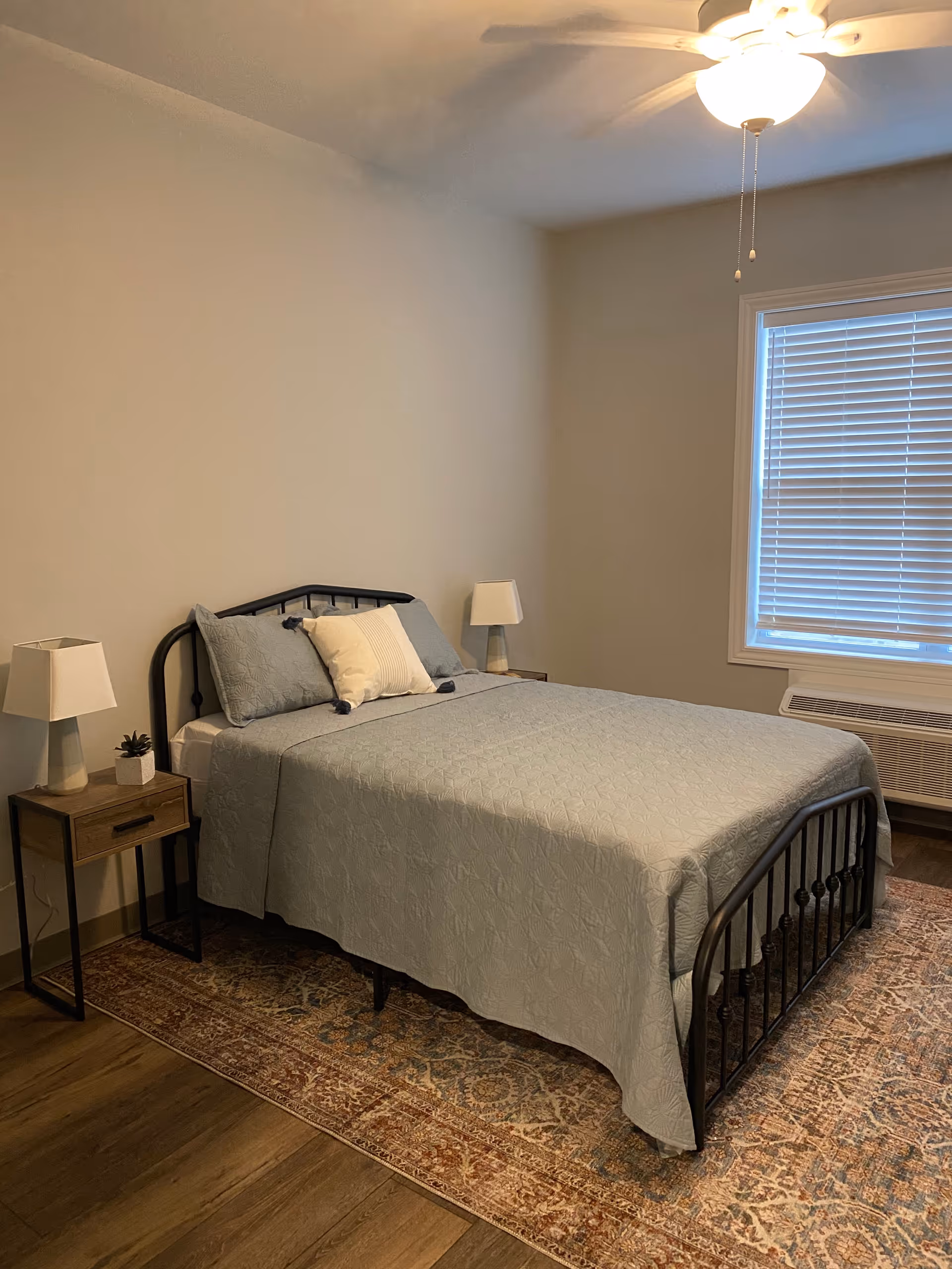 Neat bedroom with a metal-frame bed made up in blue-gray bedding, two bedside tables with lamps, a ceiling fan, window with blinds, and a patterned area rug.