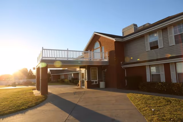 Exterior view of a senior living facility building with a covered entrance, brick and siding walls, multiple windows, and a balcony above the entrance. The sun is low in the sky, casting long shadows on the driveway and lawn.