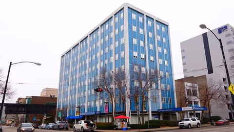 A blue glass high-rise building on a downtown street with parked cars and leafless trees