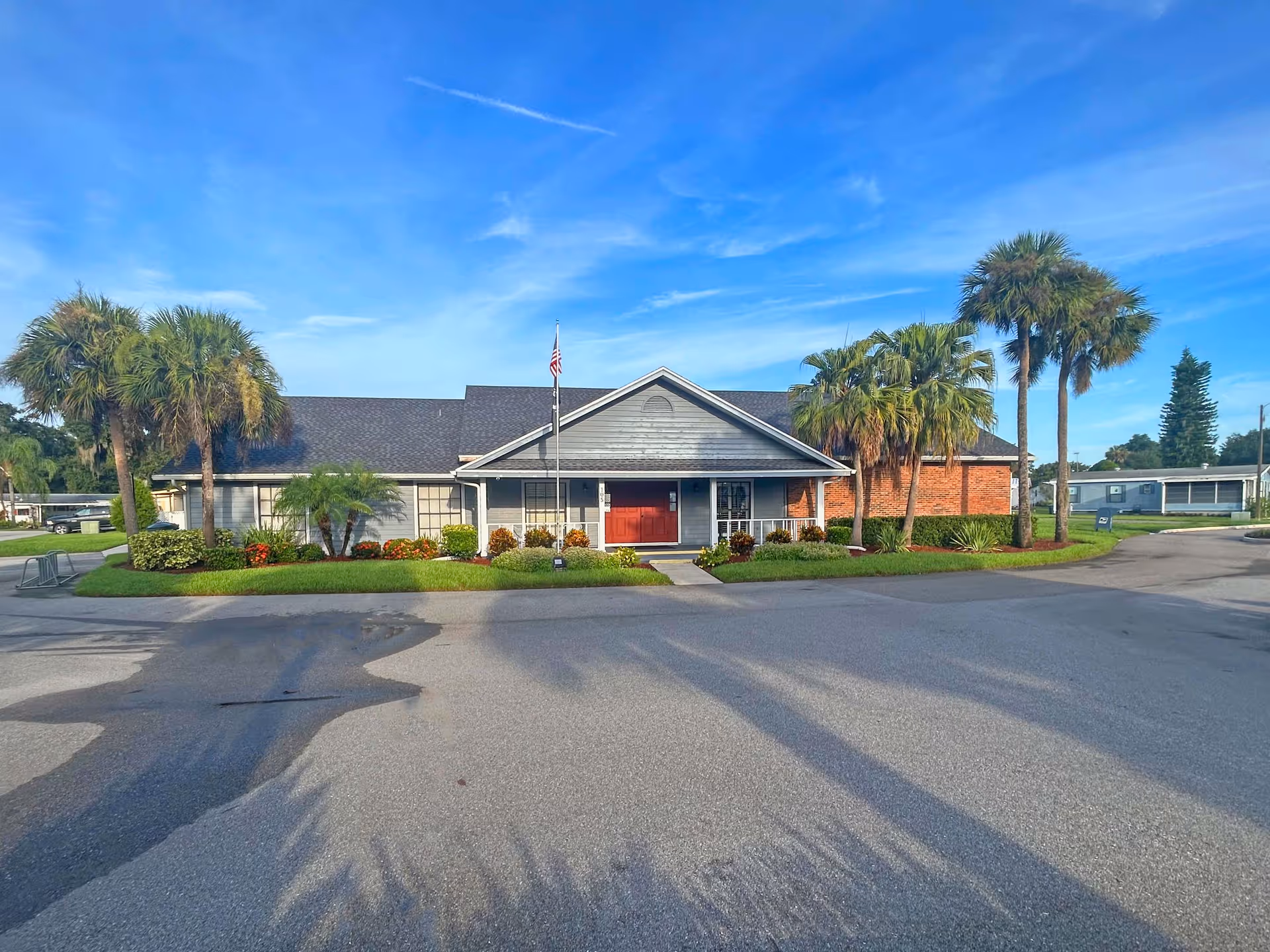 Single-story retirement community building with a flagpole, palm trees and landscaped lawn under a blue sky.