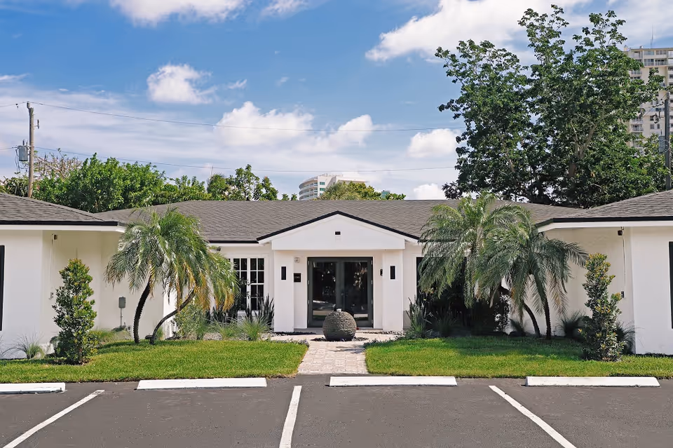 White, single-story building with a central entrance, palm trees, and a parking lot.