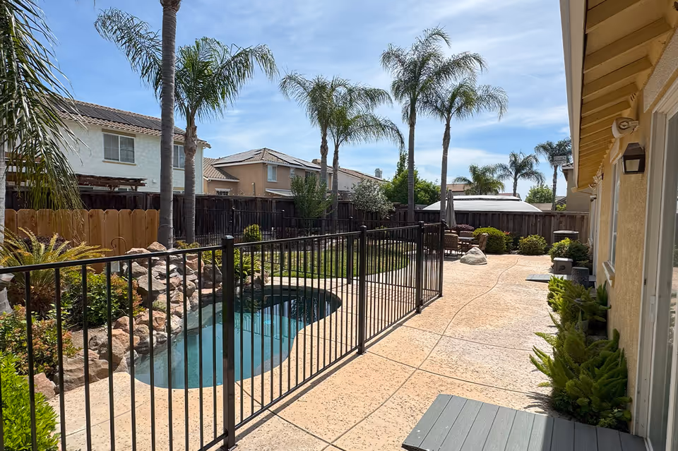 Outdoor fenced pool area with palm trees, railing, and patio furniture beside a yellow building.