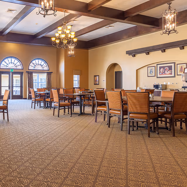 Spacious dining room with multiple wooden tables and chairs arranged neatly. The room features patterned carpet flooring, beige walls, and decorative chandeliers hanging from a ceiling with exposed wooden beams. Large arched windows and doors allow natural light to enter the room. Artwork is displayed on the walls, and there is a counter area with a coffee machine and other items.