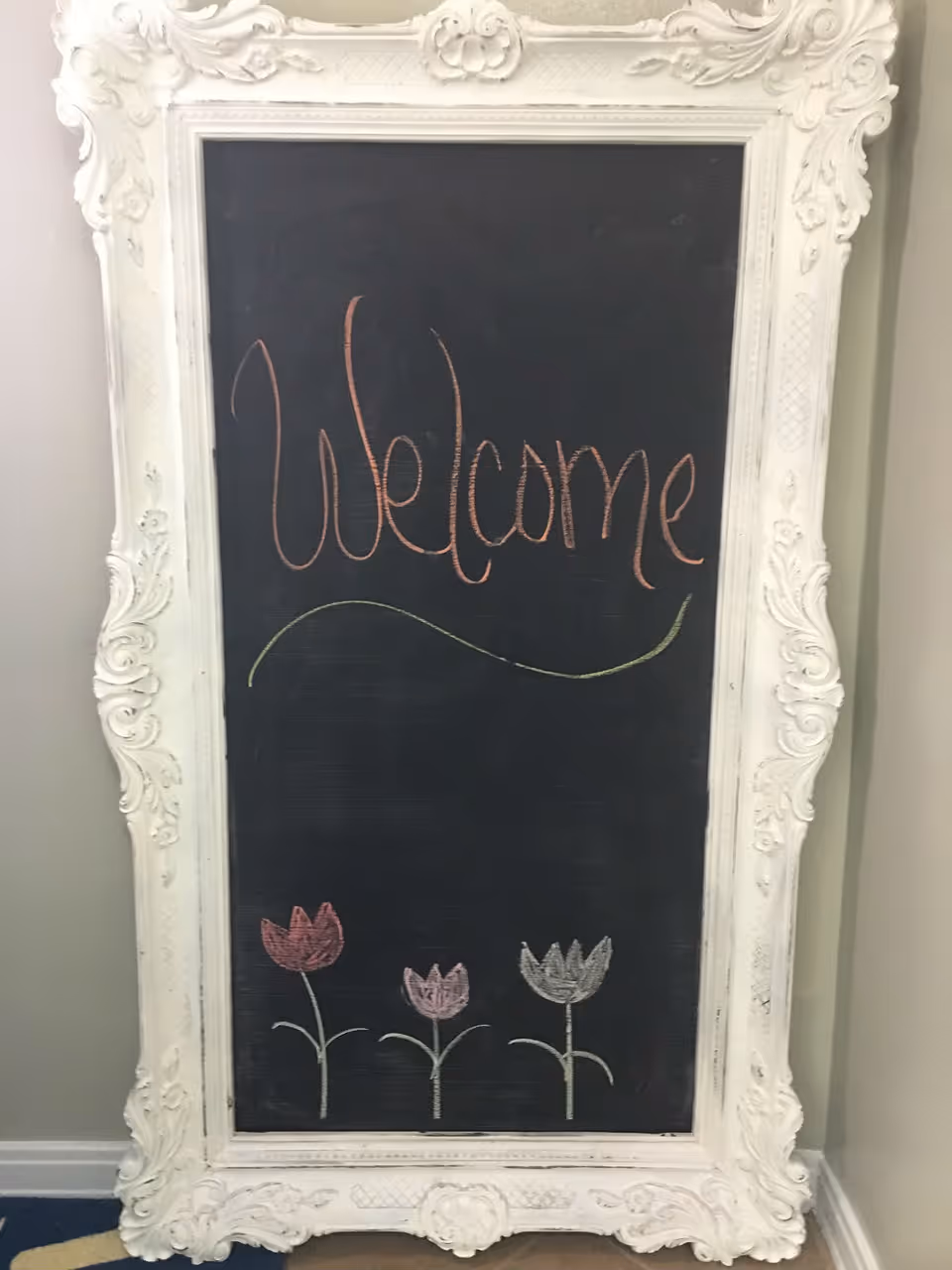 Ornate white-framed chalkboard in interior reads 'Welcome' with illustrated flowers.