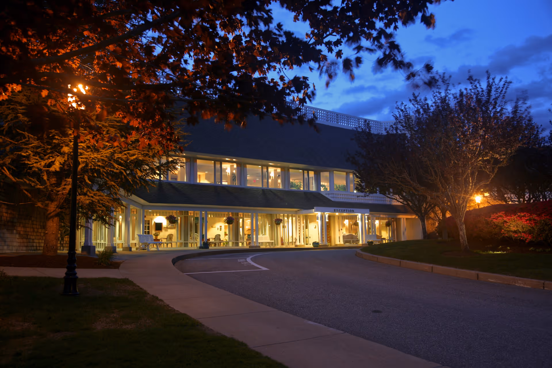 Evening view of the entrance to Thirwood Place facility with warm lights glowing from inside, surrounded by trees and a curved driveway leading to the reception area.