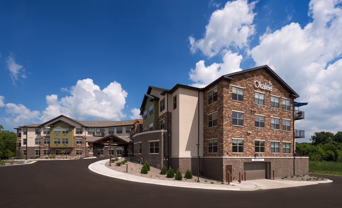 Exterior view of the Okalee of Medina multi-story senior living building with brick and stucco facades and a curved driveway under a blue sky.