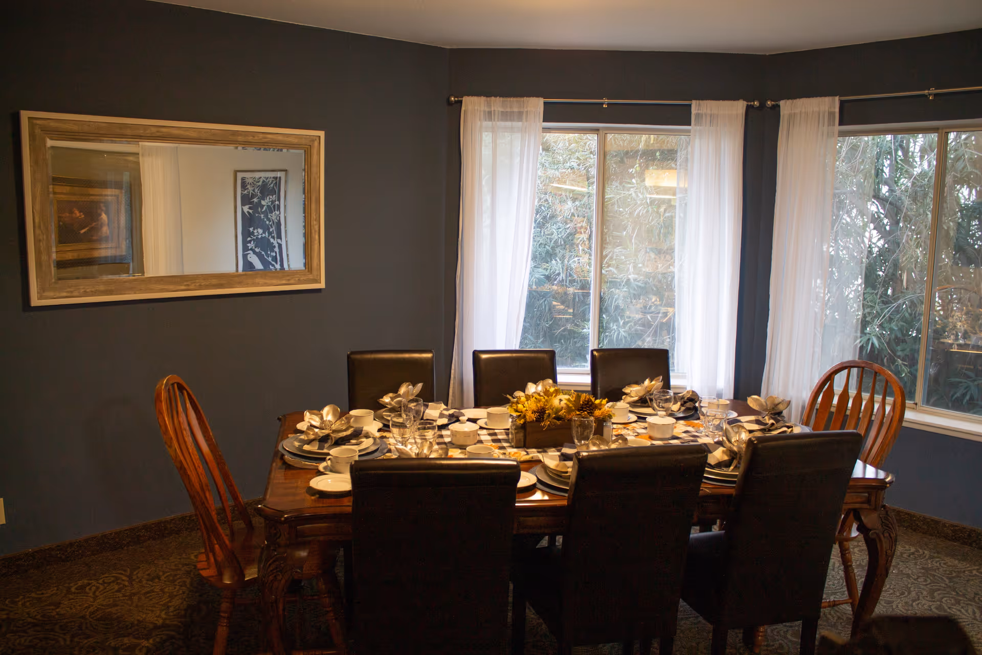 A dining room with a wooden table set for six people. The table is decorated with plates, cups, napkins, glasses, and a centerpiece with flowers. There are six chairs around the table, four with black upholstery and two wooden chairs. The room has dark blue walls, two large windows with sheer white curtains, and a large rectangular mirror on one wall.